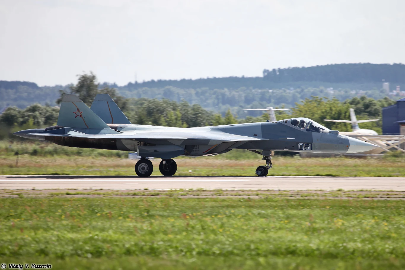 Myśliwiec wielozadaniowy Su-57 na pasie startowym, widoczny z boku podczas kołowania, w tle nisko położona zieleń i porozrzucane budynki lotniska.