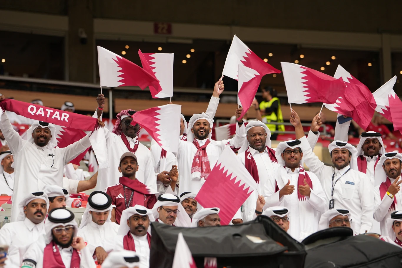 Grupa kibiców w tradycyjnych arabskich strojach ubrana w barwy narodowe Kataru, energicznie machających flagami i szalikiem z napisem „Qatar” na trybunach stadionu.