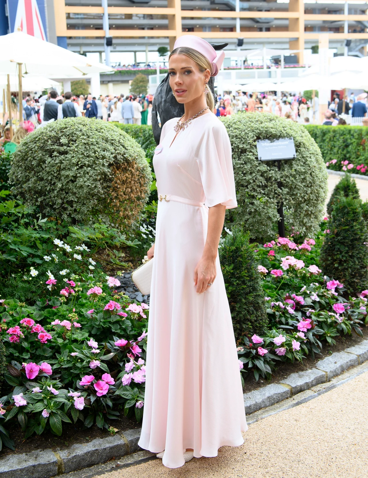 Kobieta w długiej, jasnoróżowej sukni i opasce na głowie stoi w eleganckim ogrodzie kwiatowym podczas wydarzenia towarzyskiego.