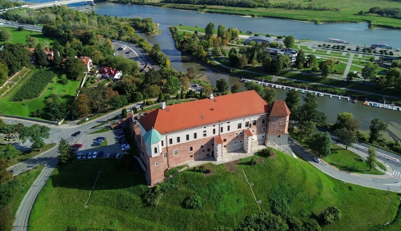 Zamek o ceglanych murach i czerwonym dachu położony na zielonym wzgórzu, otoczony krętą drogą i rzeką płynącą w tle.