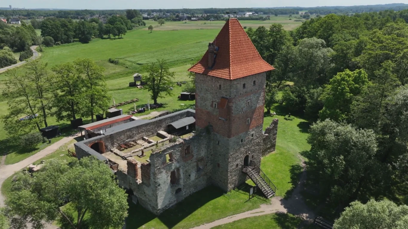 Kamienna ruina zamku z dobrze zachowaną czworoboczną wieżą i czerwonym dachem otoczona zielonymi polami i gęstym drzewostanem.