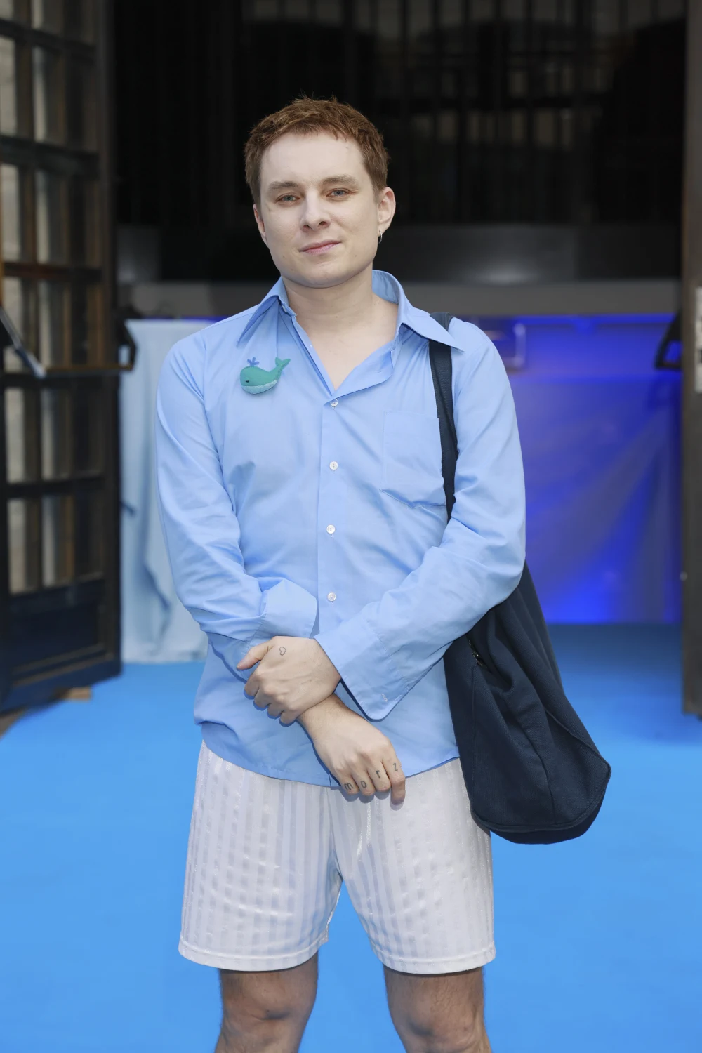 Mężczyzna w błękitnej koszuli, jasnych krótkich spodenkach i z czarną torbą na ramieniu stoi na niebieskiej podłodze, w tle otwarte drzwi i delikatne niebieskie oświetlenie.