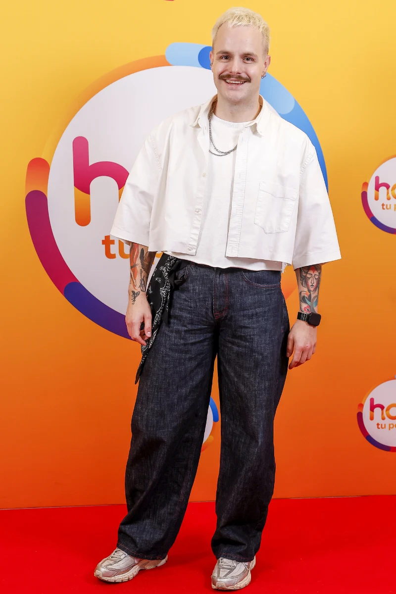 Filip Mettler w "halo tu polsat" Uśmiechnięta osoba z jasnymi włosami ubrana w białą, krótką koszulę, luźną białą koszulkę, szerokie ciemne spodnie dżinsowe i sportowe buty, z widocznymi tatuażami na rękach, stojąca na czerwonym dywanie na tle pomarańczowej ścianki z logotypem.