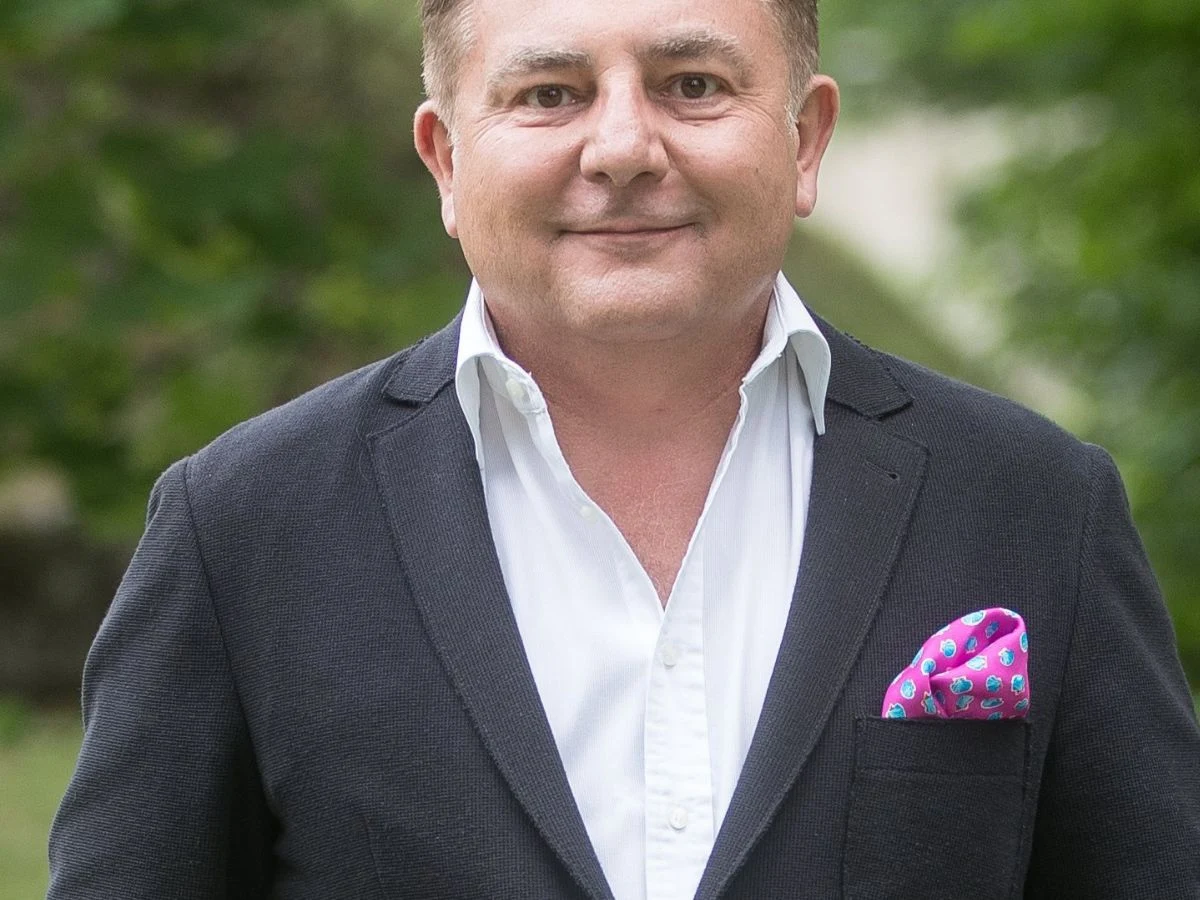 Robert Makłowicz Robert Makłowicz w ciemnej marynarce z różową poszetką w kieszonce, pozujący na tle zieleni.