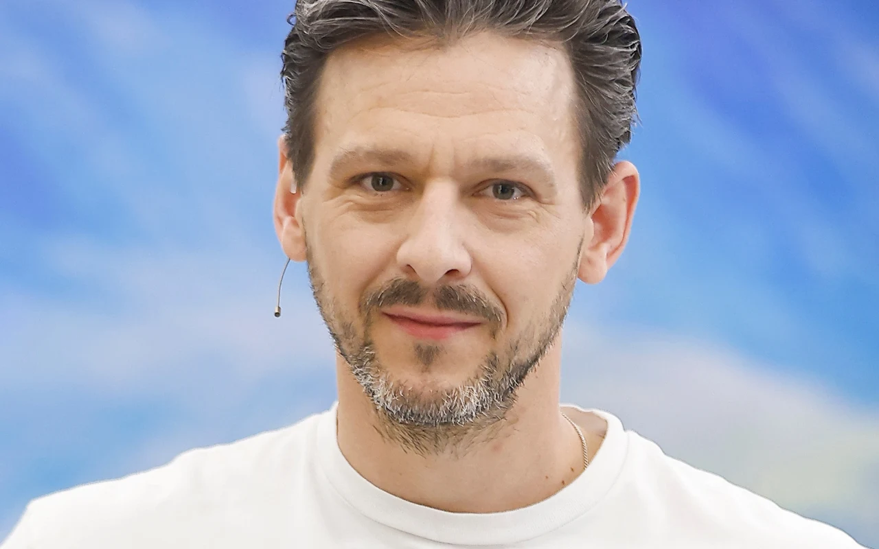 Mateusz Kmiecik Mateusz Kmiecik, ubrany w biały T-shirt, na tle niebieskiego, rozmytego tła.