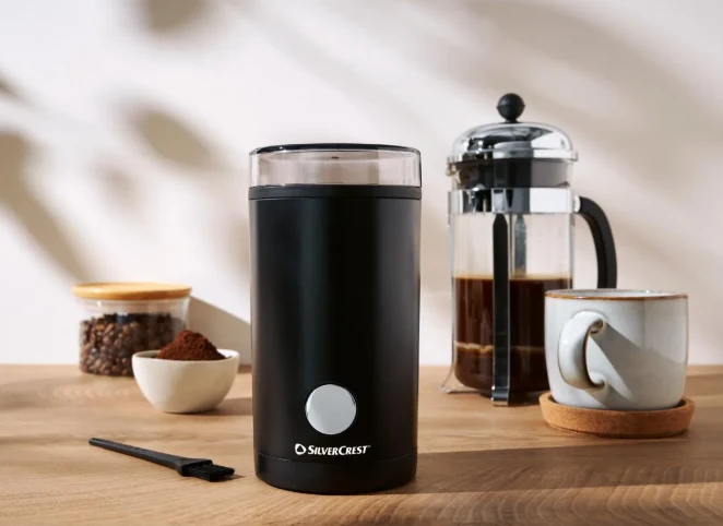 Czarny młynek do kawy marki Silvercrest na drewnianym blacie, obok szklany zaparzacz French Press wypełniony kawą, filiżanka na spodku, przezroczysty słoiczek z ziarnami kawy i mała miseczka ze zmieloną kawą.