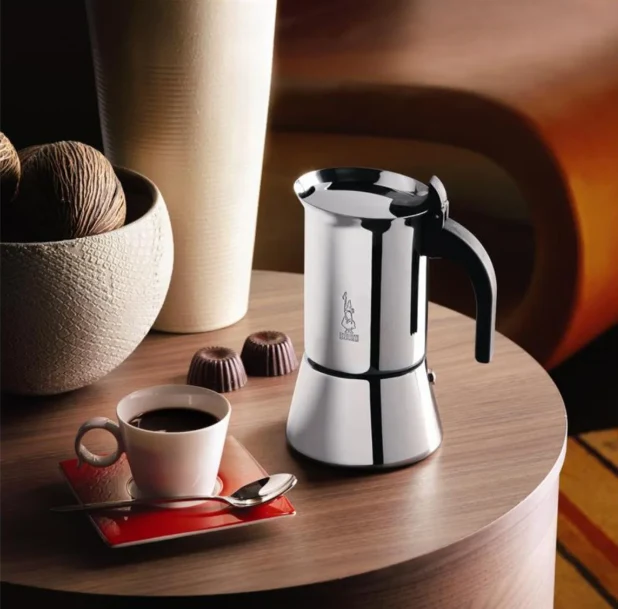 Metalowy czajniczek do kawy typu moka ustawiony obok filiżanki espresso na czerwonym spodku oraz dwóch czekoladek na okrągłym drewnianym stoliku, w tle dekoracyjna misa z kłębkami sznurka.