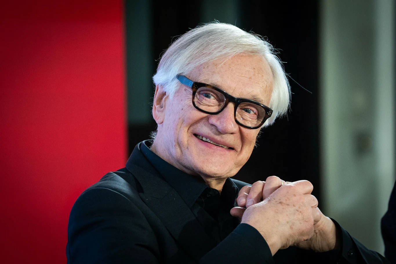 Starszy mężczyzna o siwych włosach i okularach, uśmiechnięty, ubrany w ciemny strój, siedzi z dłońmi splecionymi przed sobą na tle czerwonej ściany.
