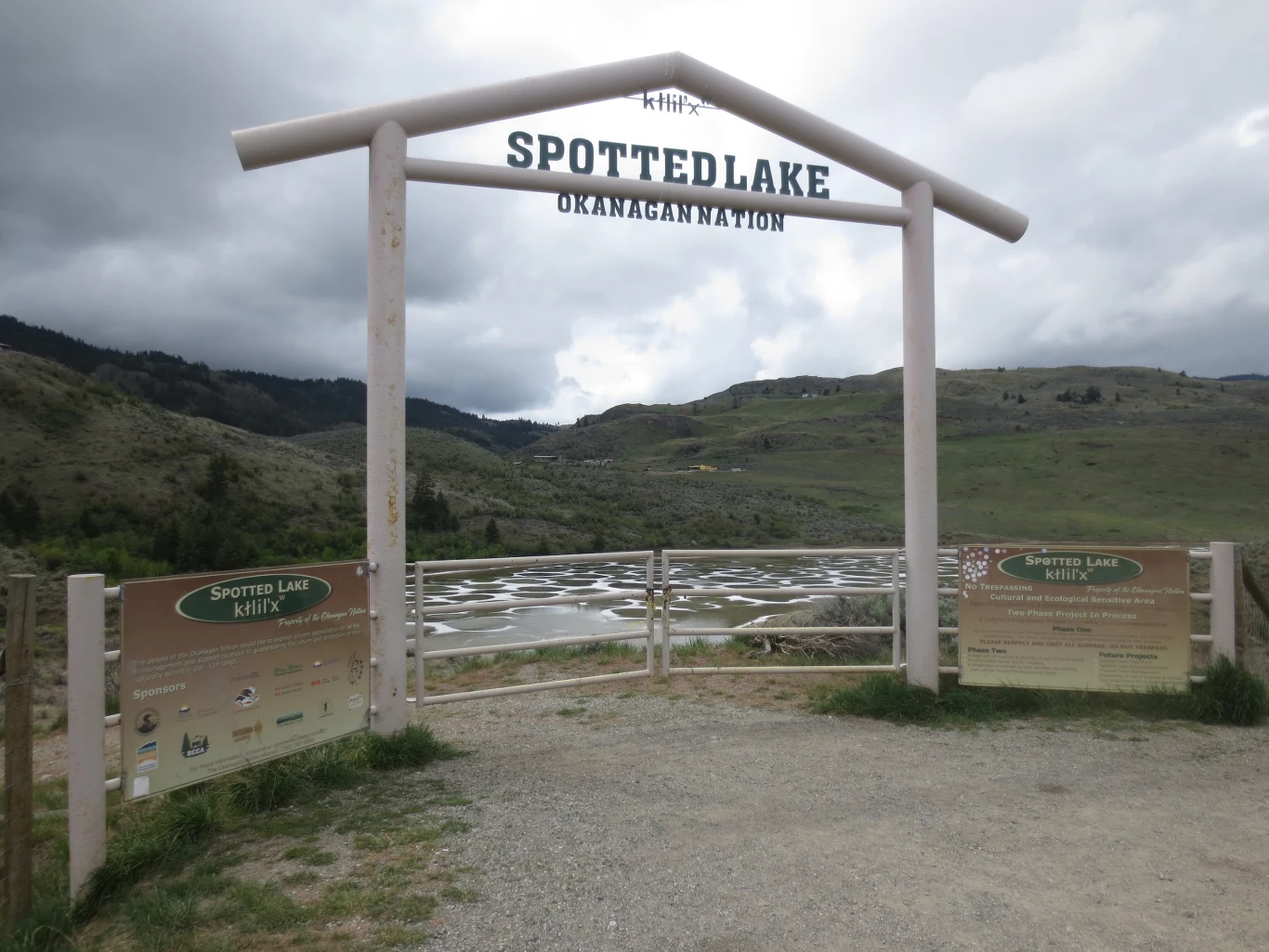 drewniana brama z napisem „Spotted Lake Okanagan Nation” stojąca przed charakterystycznym, nakrapianym jeziorem otoczonym pagórkowatym, skalistym krajobrazem