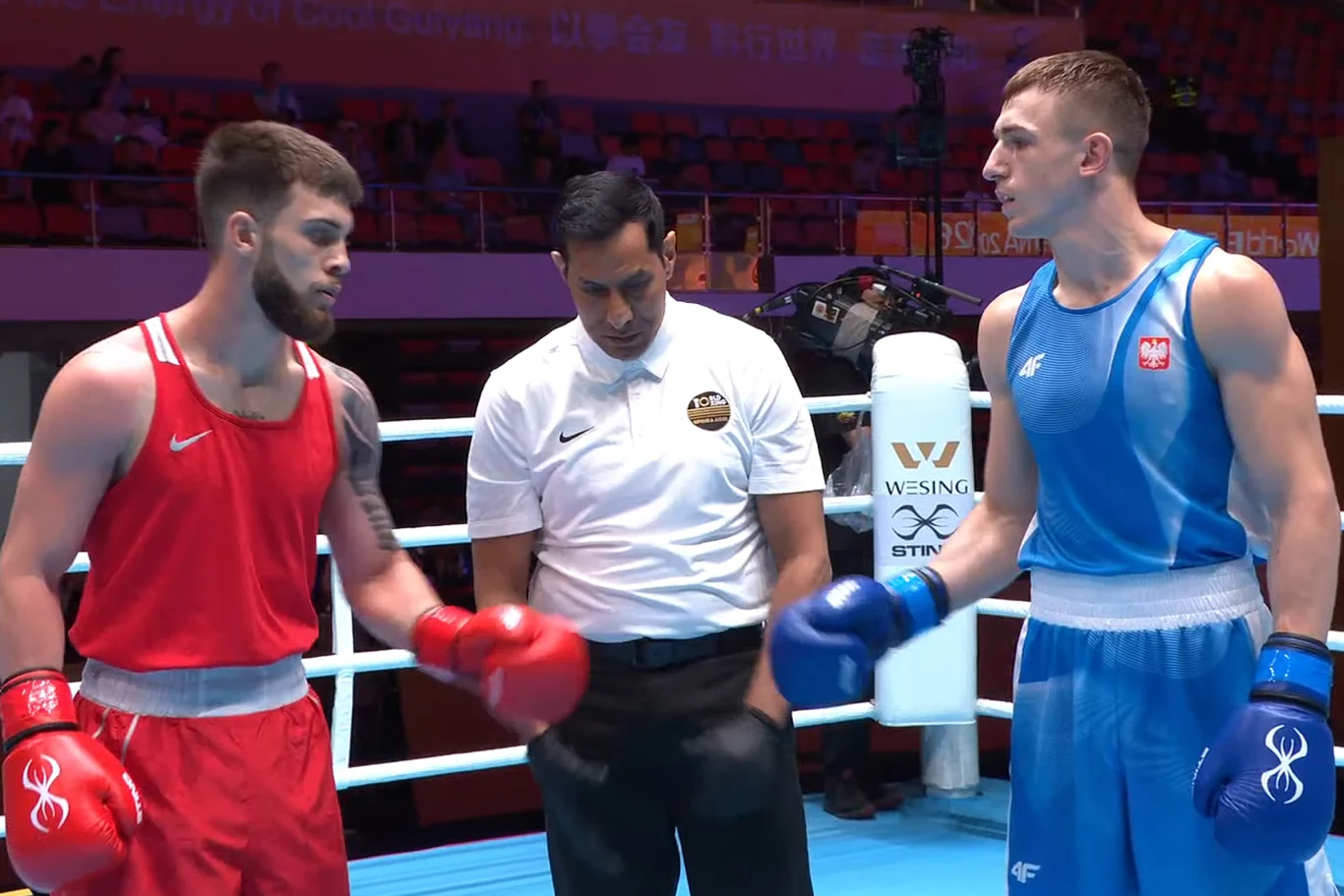 Giorgi Natroszwili i Mateusz Urban przed walką w Guiyangu Dwóch bokserów w strojach sportowych, jeden w czerwonym, drugi w niebieskim, stoi naprzeciwko siebie na ringu, między nimi arbiter w białej koszulce.