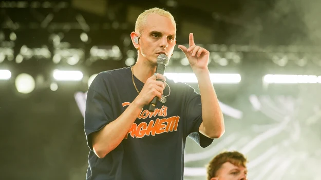 Wokalista z mikrofonem podczas koncertu, ubrany w ciemny t-shirt z dużym nadrukiem, w tle rozświetlona scena i drugi muzyk.