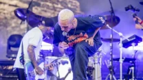 Wokalista z jasnymi włosami śpiewa do mikrofonu na scenie koncertowej, towarzyszy mu gitarzysta z tatuażami na rękach, w tle widoczne są perkusja i profesjonalny sprzęt muzyczny.