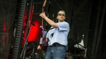 Mężczyzna w okularach przeciwsłonecznych grający na gitarze elektrycznej na scenie koncertowej, skąpanej w czerwonym świetle scenicznym.