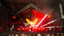 Zespół muzyczny występujący na dużej scenie podczas koncertu, efekty pirotechniczne w postaci wybuchu ognia, intensywne oświetlenie sceniczne oraz tłum publiczności obserwujący wydarzenie z bliska.