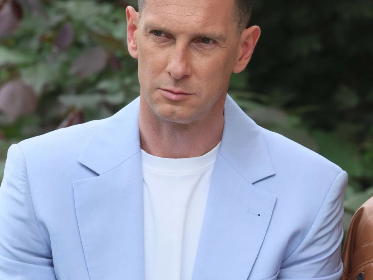 Łukasz Kadziewicz Łukasz Kadziewicz w jasnoniebieskiej marynarce i białej koszulce na tle zieleni.