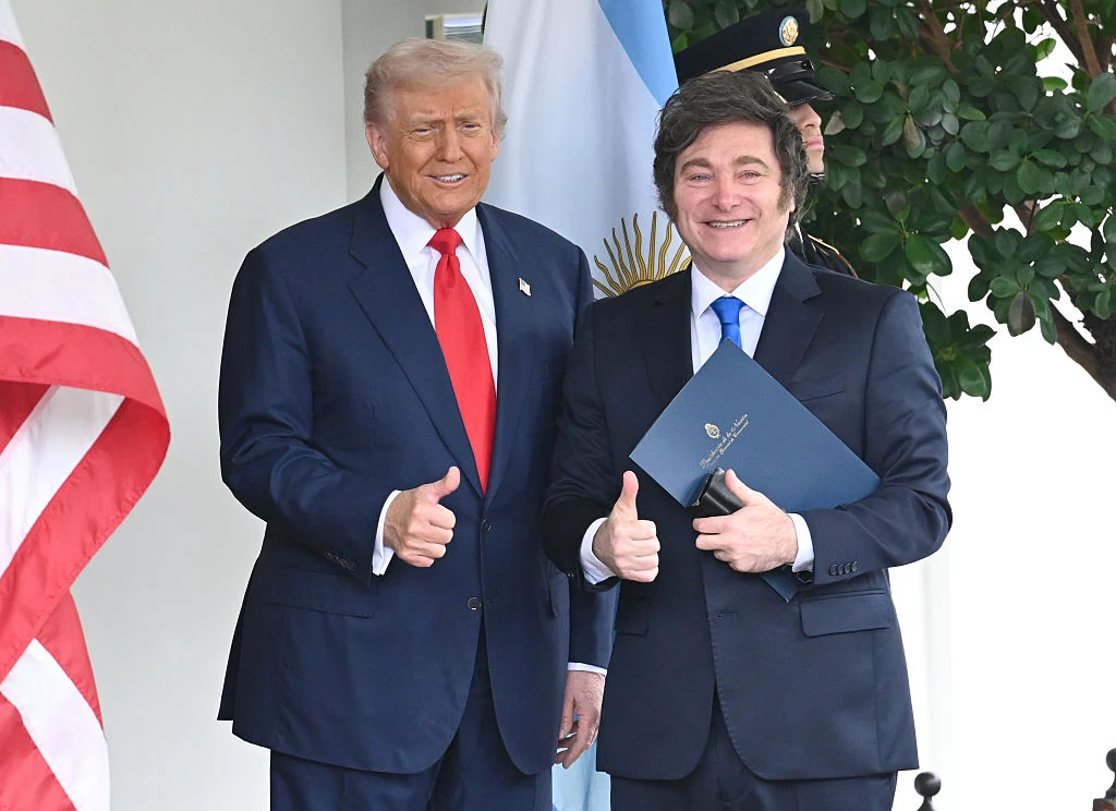 Prezydenci USA i Argentyny: Donald Trump oraz Javier Milei Dwóch mężczyzn w garniturach stoi obok siebie, obaj uśmiechają się i pokazują kciuki uniesione w górę, w tle widoczne są flagi Stanów Zjednoczonych i Argentyny oraz roślina doniczkowa.