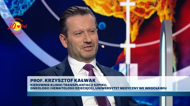 Mężczyzna w garniturze i krawacie z mikrofonem przy twarzy udziela wywiadu w studiu telewizyjnym; na ekranie wyświetlone jest jego imię, nazwisko oraz tytuł naukowy i stanowisko zawodowe.
