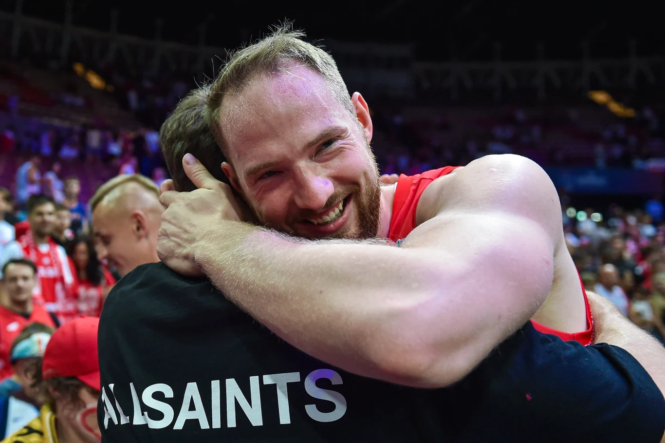 Uśmiechnięty mężczyzna w stroju sportowym obejmuje drugą osobę w tłumie na trybunach podczas wydarzenia sportowego.