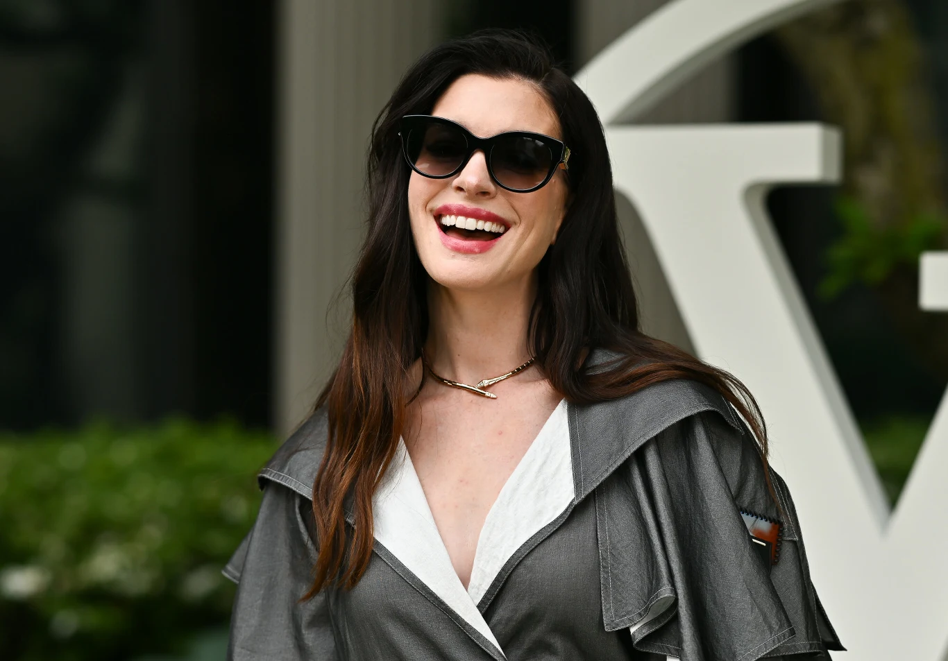Anne Hathaway Anne Hathaway w eleganckim szarym płaszczu i okularach przeciwsłonecznych, na tle białej architektury.
