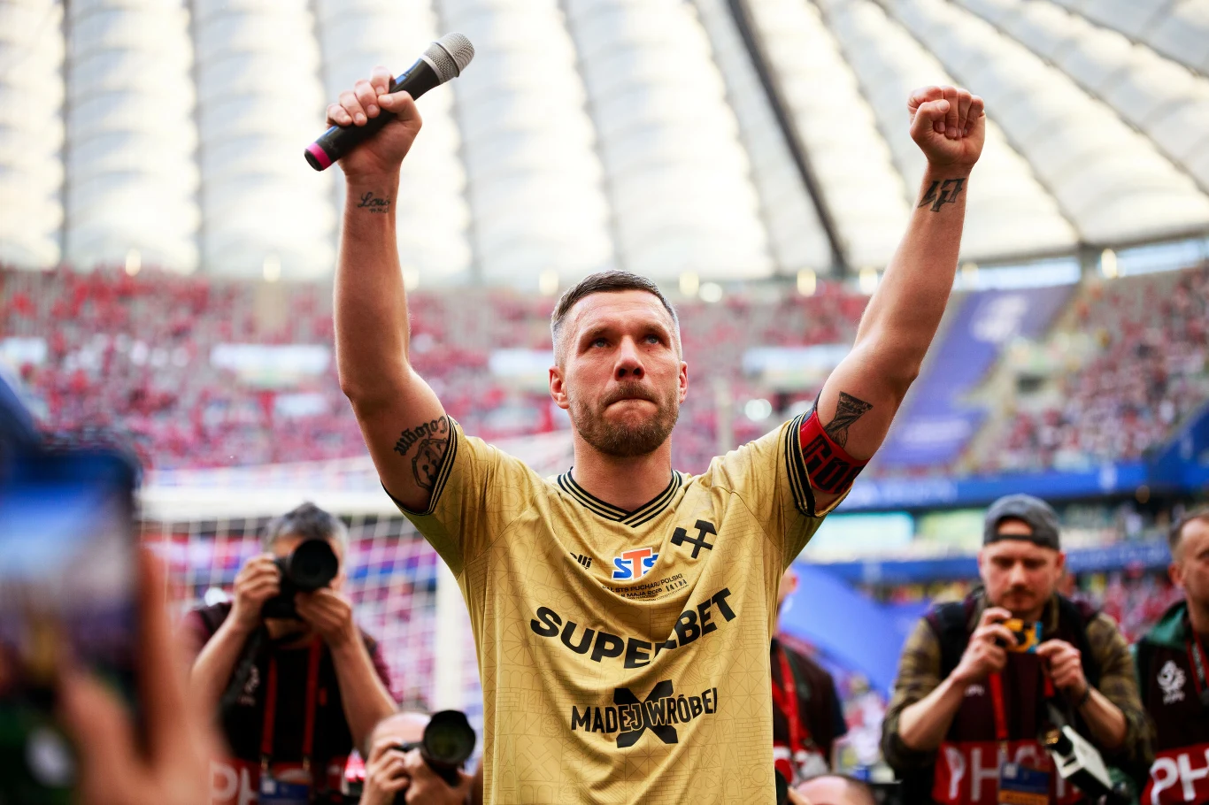 Piłkarz w złotej koszulce z opaską kapitańską unosi jedną rękę w geście triumfu i trzyma mikrofon, otoczony przez fotografów na stadionie pełnym kibiców.