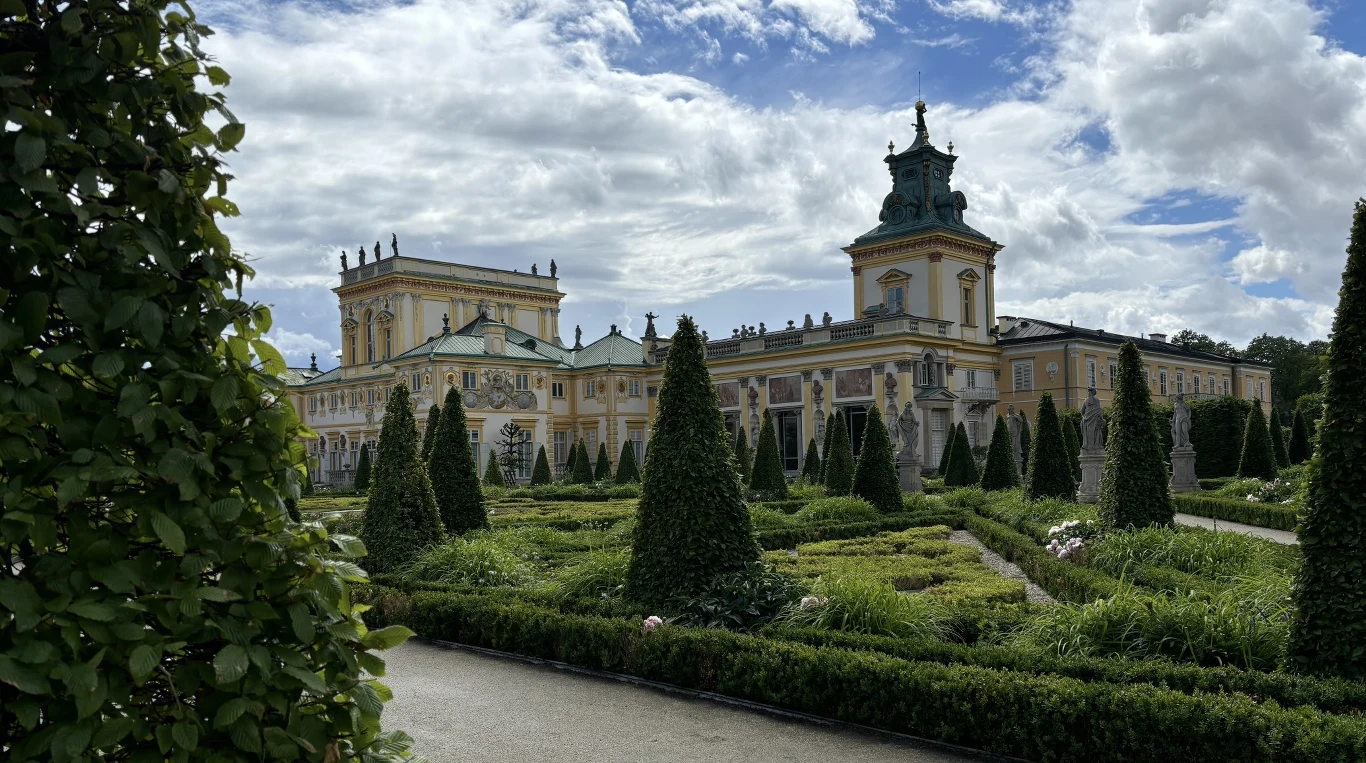 Barokowy pałac z wysoką wieżą otoczony symetrycznym ogrodem z równo przyciętymi żywopłotami i stożkowymi krzewami pod jasnym, lekko zachmurzonym niebem.