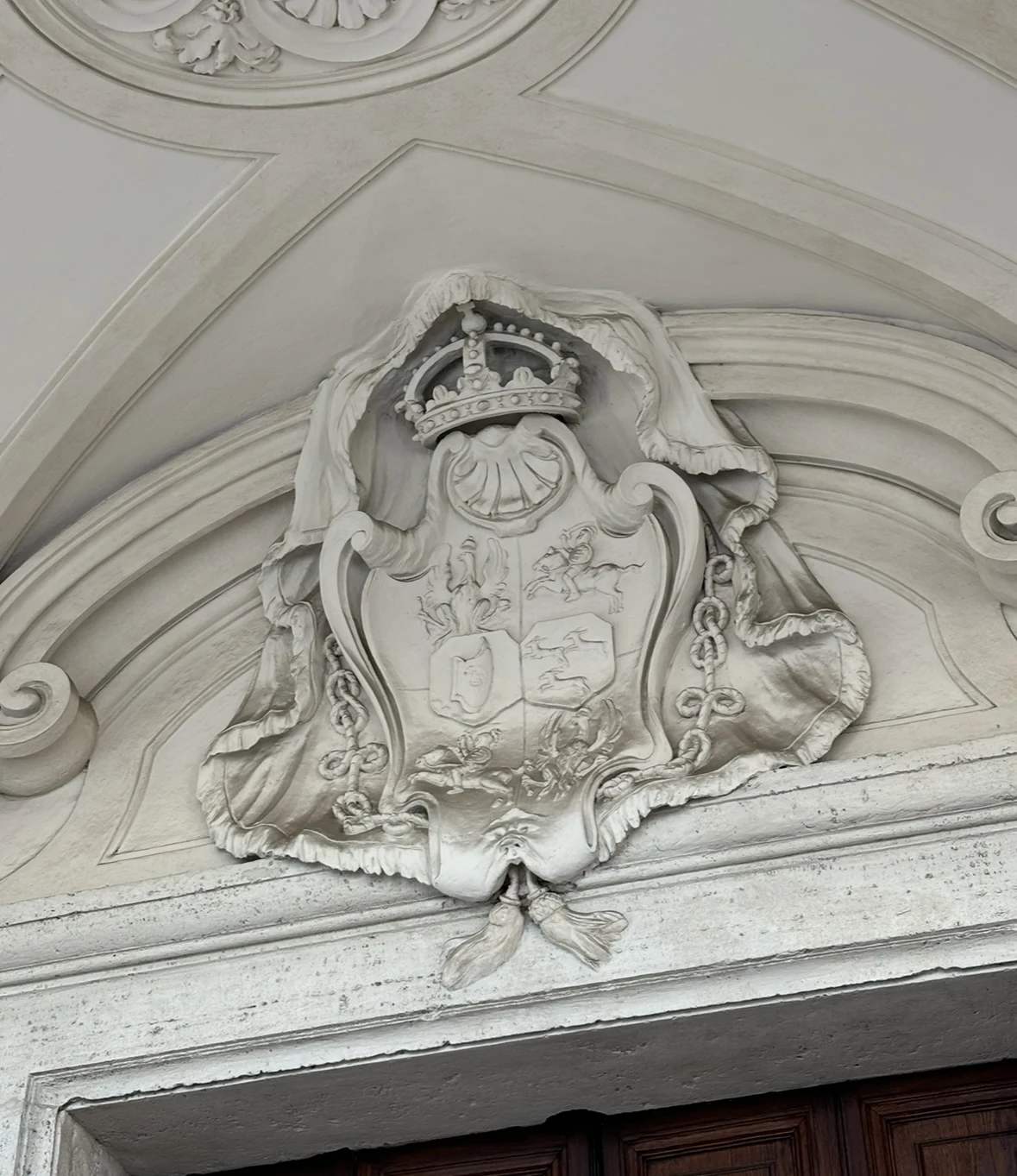 Kamienny herb umieszczony nad wejściem, bogato zdobiony koroną, motywami roślinnymi i symbolami heraldycznymi, podtrzymywany przez fałdy szaty ornamentalnej.