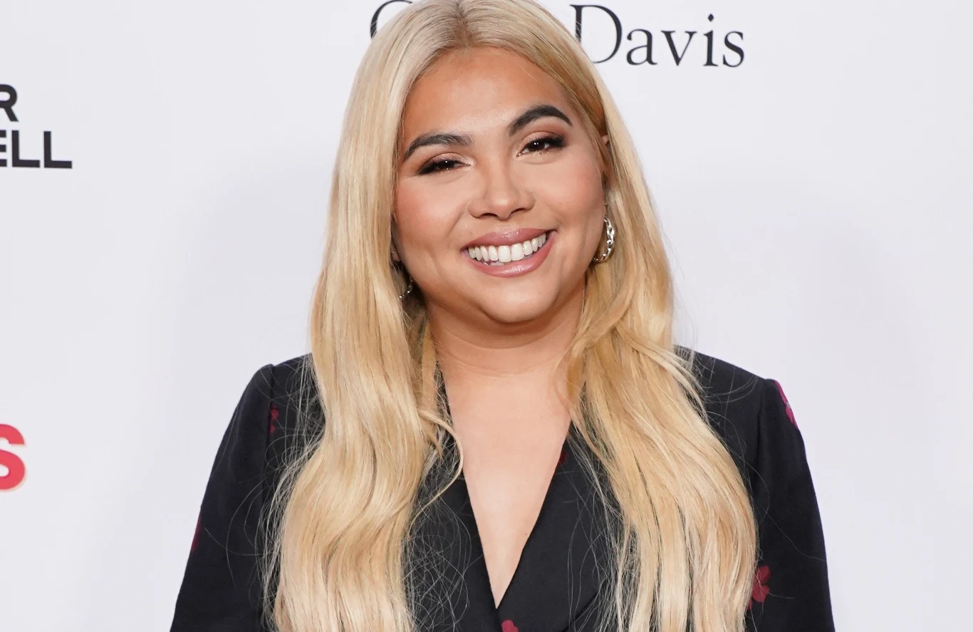Hayley Kiyoko Hayley Kiyoko z długimi blond włosami, w czarnej marynarce, stoi na jasnym tle z fragmentami napisów.