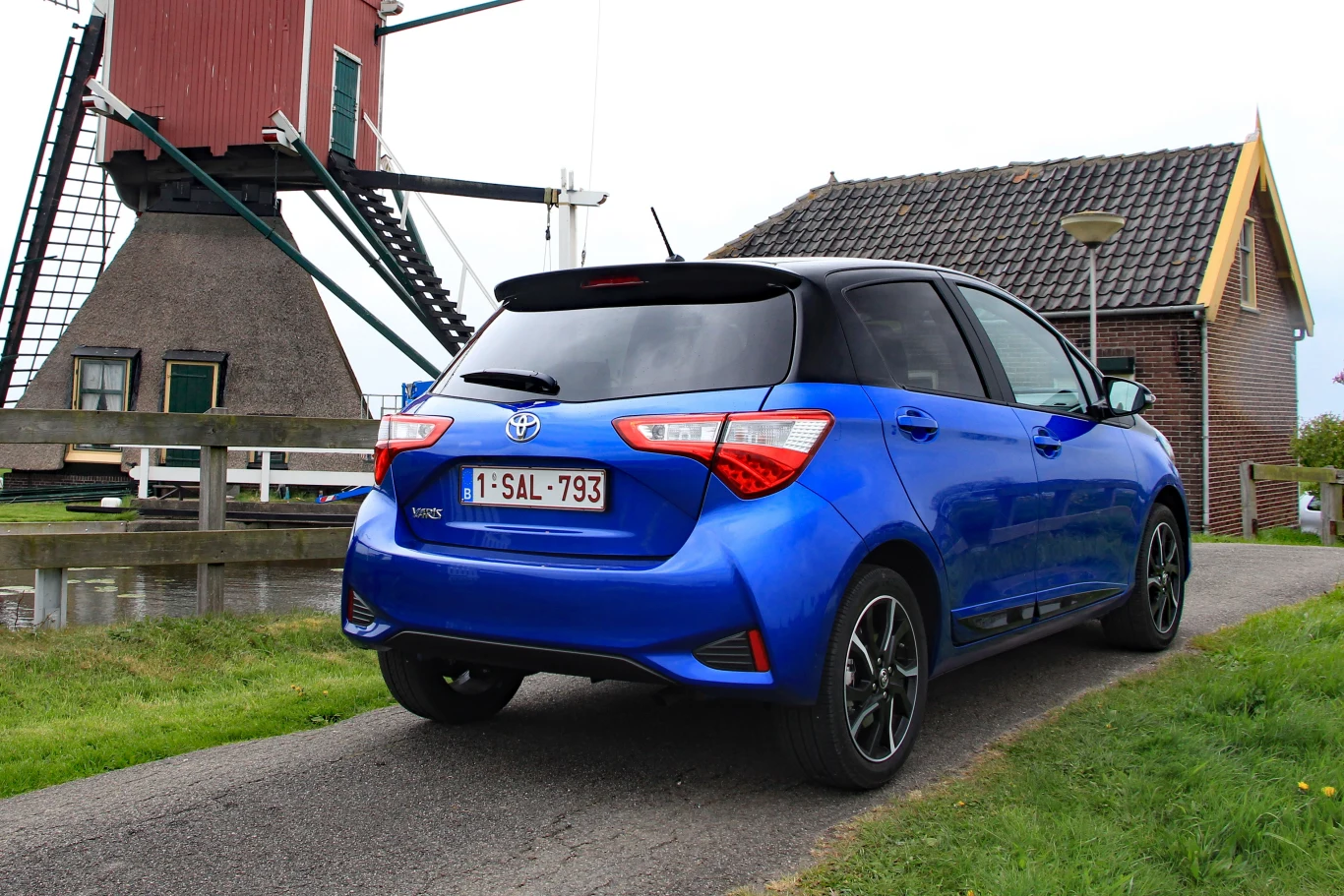 Niebieska Toyota Yaris zaparkowana na wąskiej asfaltowej drodze, w sąsiedztwie tradycyjnego holenderskiego wiatraka i niewielkiego budynku z cegły i dachówką.