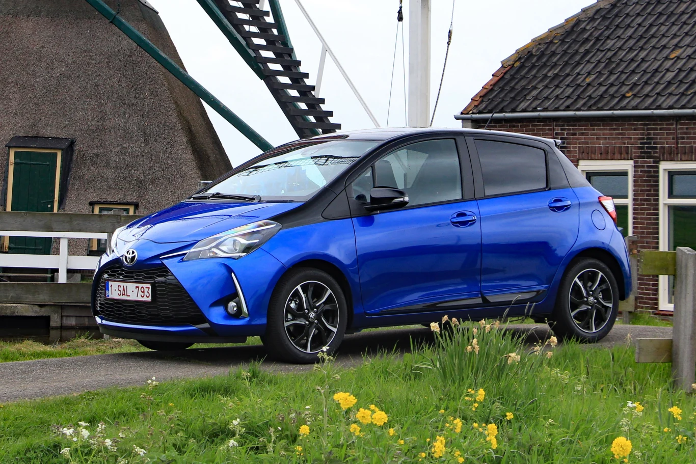 Niebieska Toyota Yaris na czarnych felgach zaparkowana przy wiejskiej drodze, w tle fragment tradycyjnego młyna i budynek z ceglanym dachem.