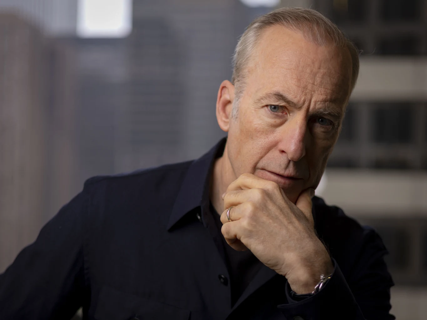 Bob Odenkirk Bob Odenkirk wspierający twarz na dłoni, w ciemnej koszuli, z pierścionkiem, na tle miejskiej zabudowy za oknem.