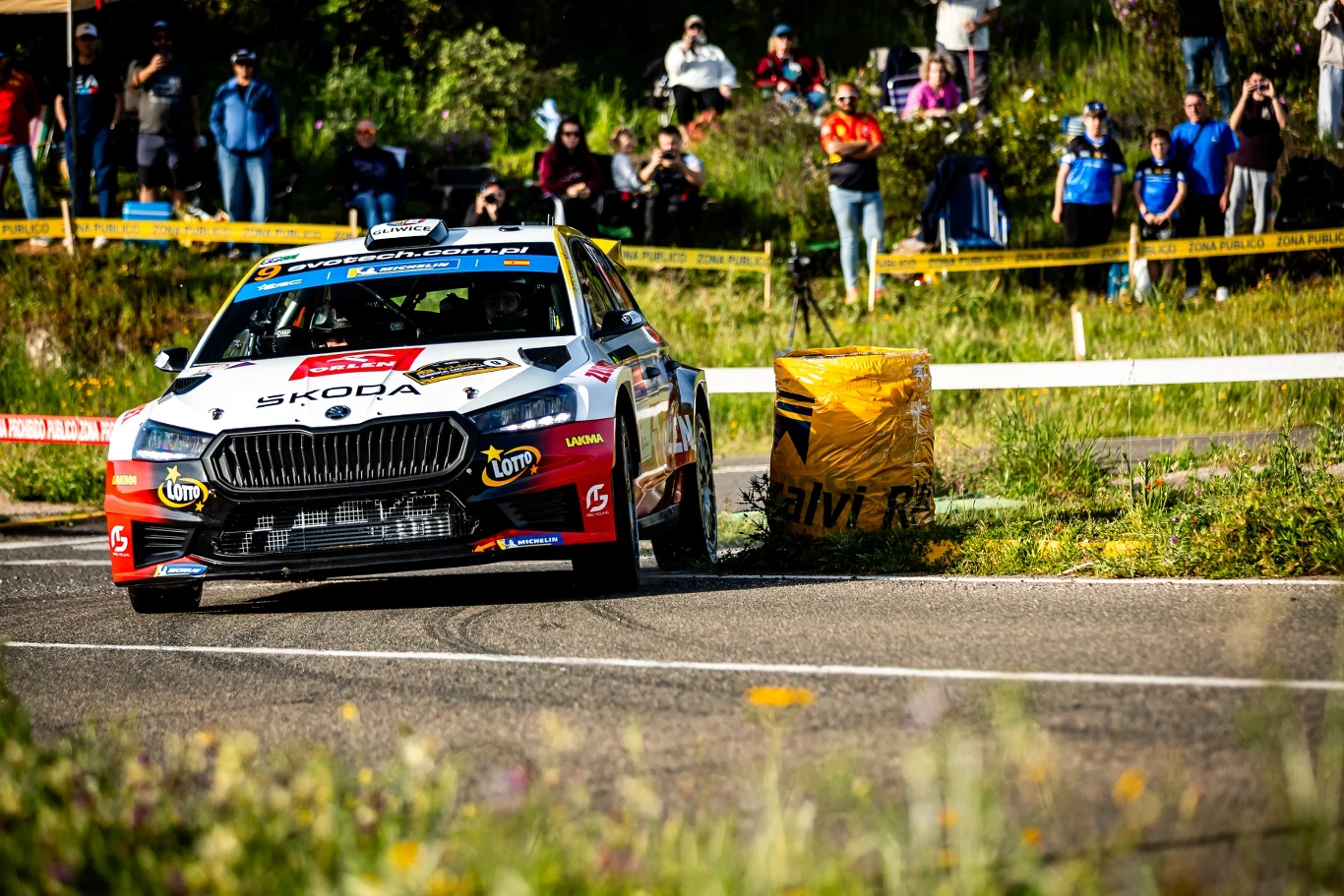 Jakub Matulka w akcji. Samochód rajdowy Škoda z numerem startowym dynamicznie pokonuje zakręt na asfaltowej trasie, mijając żółty balisad, za którym stoją kibice obserwujący wyścig.