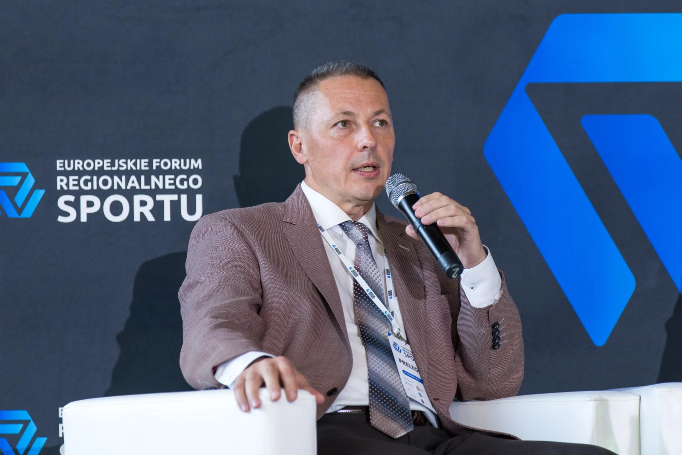 Mężczyzna w marynarce i krawacie siedzi na białym fotelu, trzymając mikrofon i przemawiając podczas wydarzenia Europejskiego Forum Regionalnego Sportu na tle czarnej ścianki z logo.