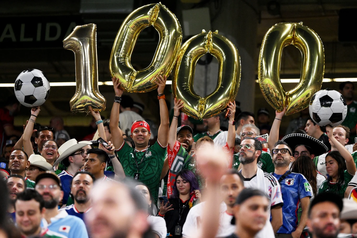 Grupa kibiców ubranych w koszulki piłkarskie trzymających duże złote balony tworzące liczbę 1000 oraz piłki futbolowe na stadionie podczas meczu.