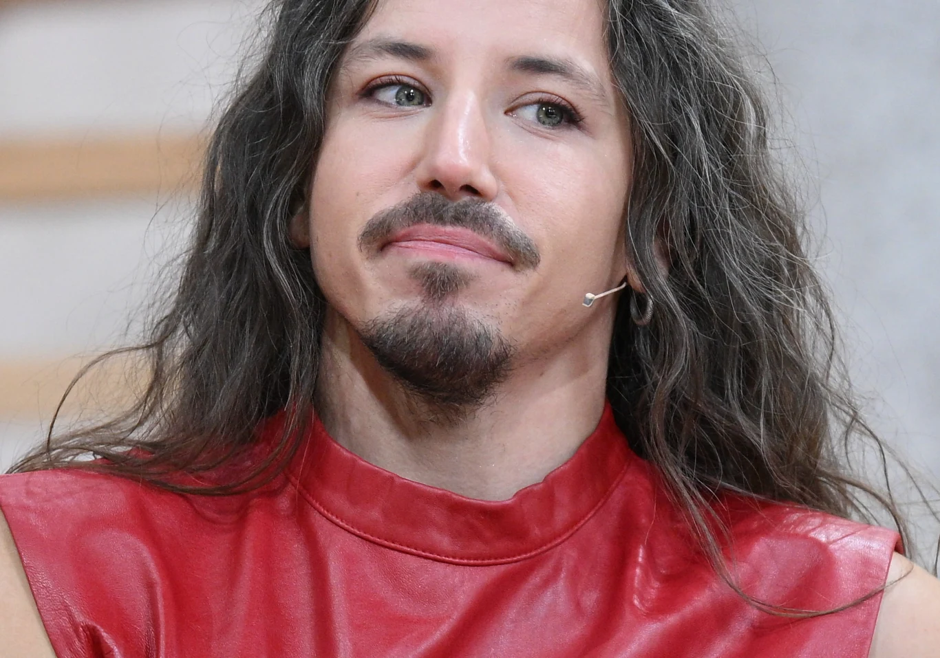 Michał Szpak Michał Szpak z długimi włosami i zarostem, ubrany w czerwoną kamizelkę, z mikrofonem przy ustach.