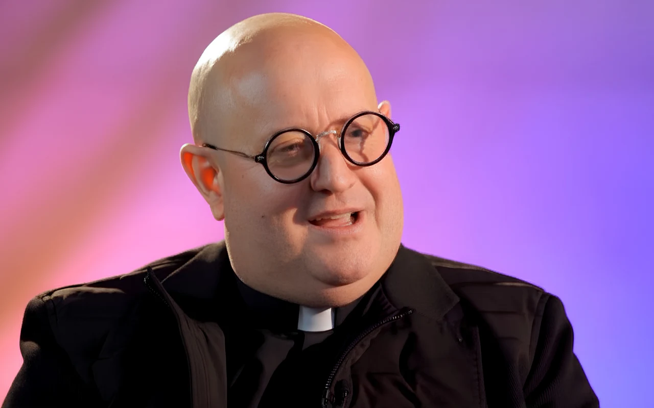 Padre Guilherme w rozmowie z Kamilą Ryciak Łysy mężczyzna w koloratce i okrągłych okularach uśmiecha się podczas rozmowy, ubrany w czarną marynarkę na kolorowym tle.