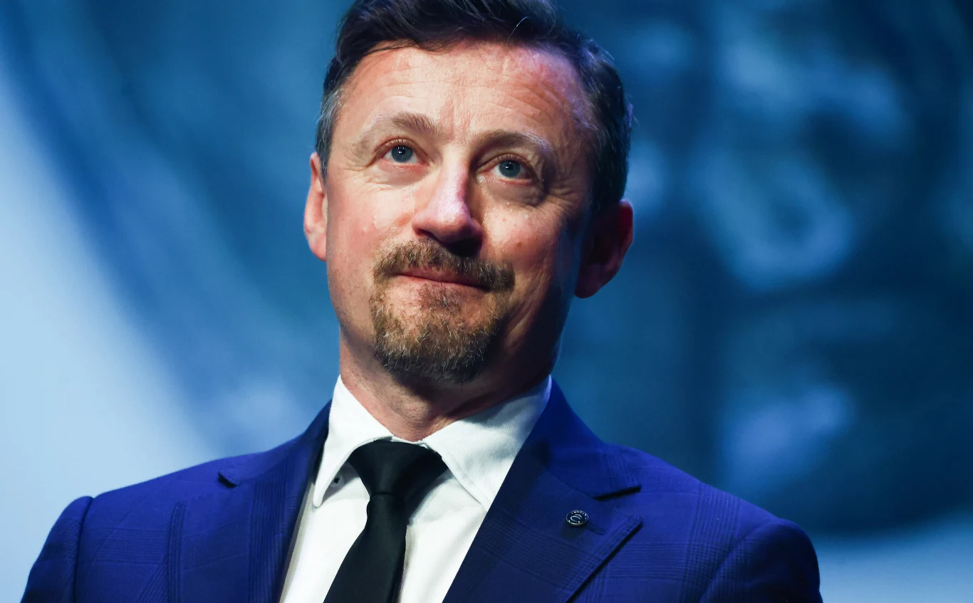 Adam Małysz Adam Małysz w granatowym garniturze, białej koszuli i czarnym krawacie na niebieskim tle.