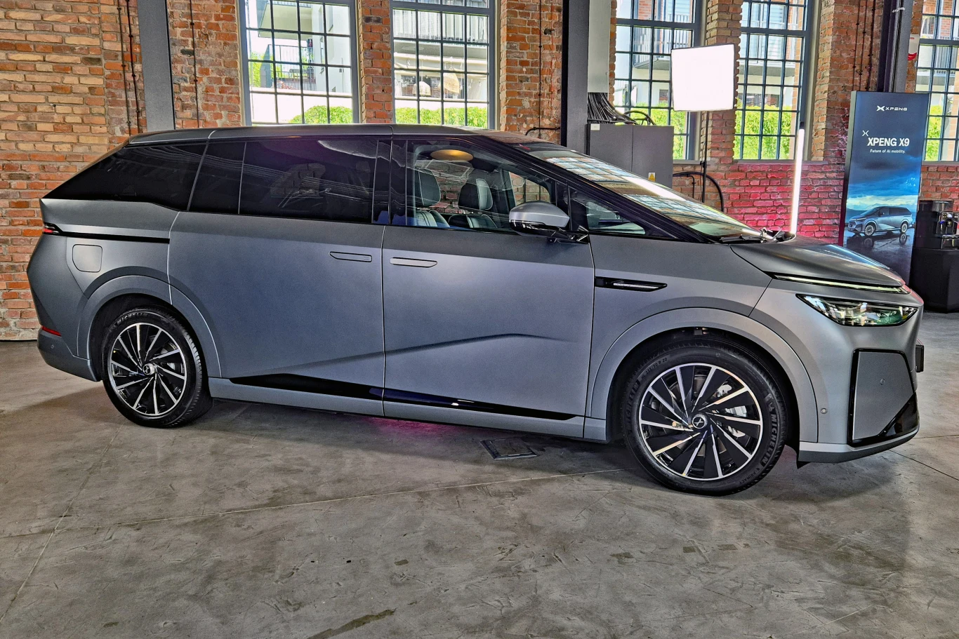 Nowoczesny, srebrnoszary minivan o dynamicznych liniach nadwozia, dużych czarnych felgach i przyciemnianych szybach, stojący w jasnej, industrialnej przestrzeni z ceglaną ścianą i przeszklonymi oknami.