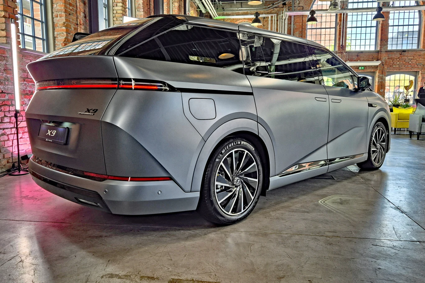 Nowoczesny minivan o aerodynamicznej sylwetce w matowym, szarym kolorze, z charakterystycznym poziomym pasem świateł LED z tyłu oraz dużymi, srebrnymi felgami na tle industrialnego wnętrza z czerwonymi cegłami i dużymi oknami.