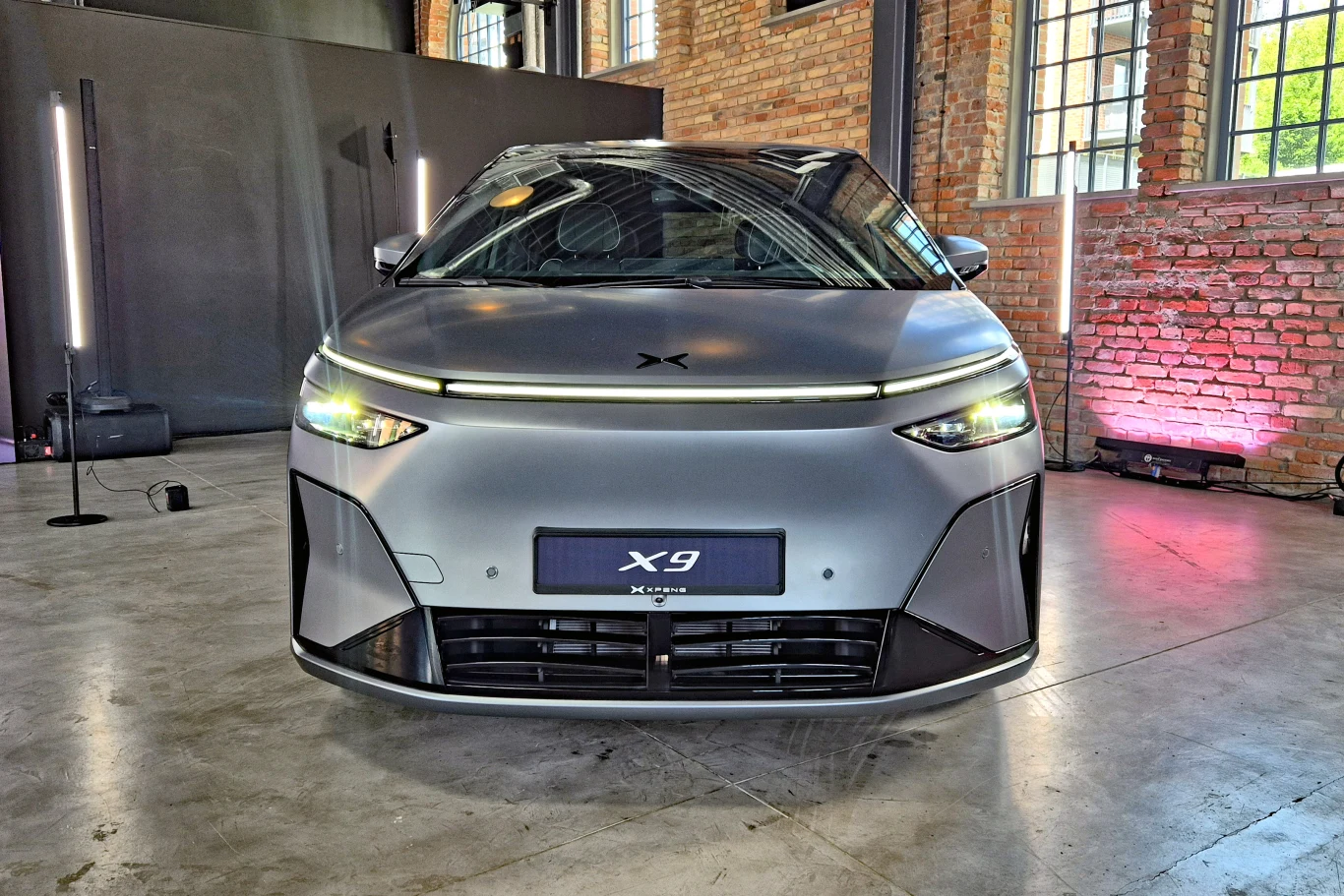 Nowoczesny elektryczny samochód Xpeng X9 w matowym, srebrnym kolorze, prezentowany w industrialnym wnętrzu z ceglanymi ścianami oraz dużymi oknami.