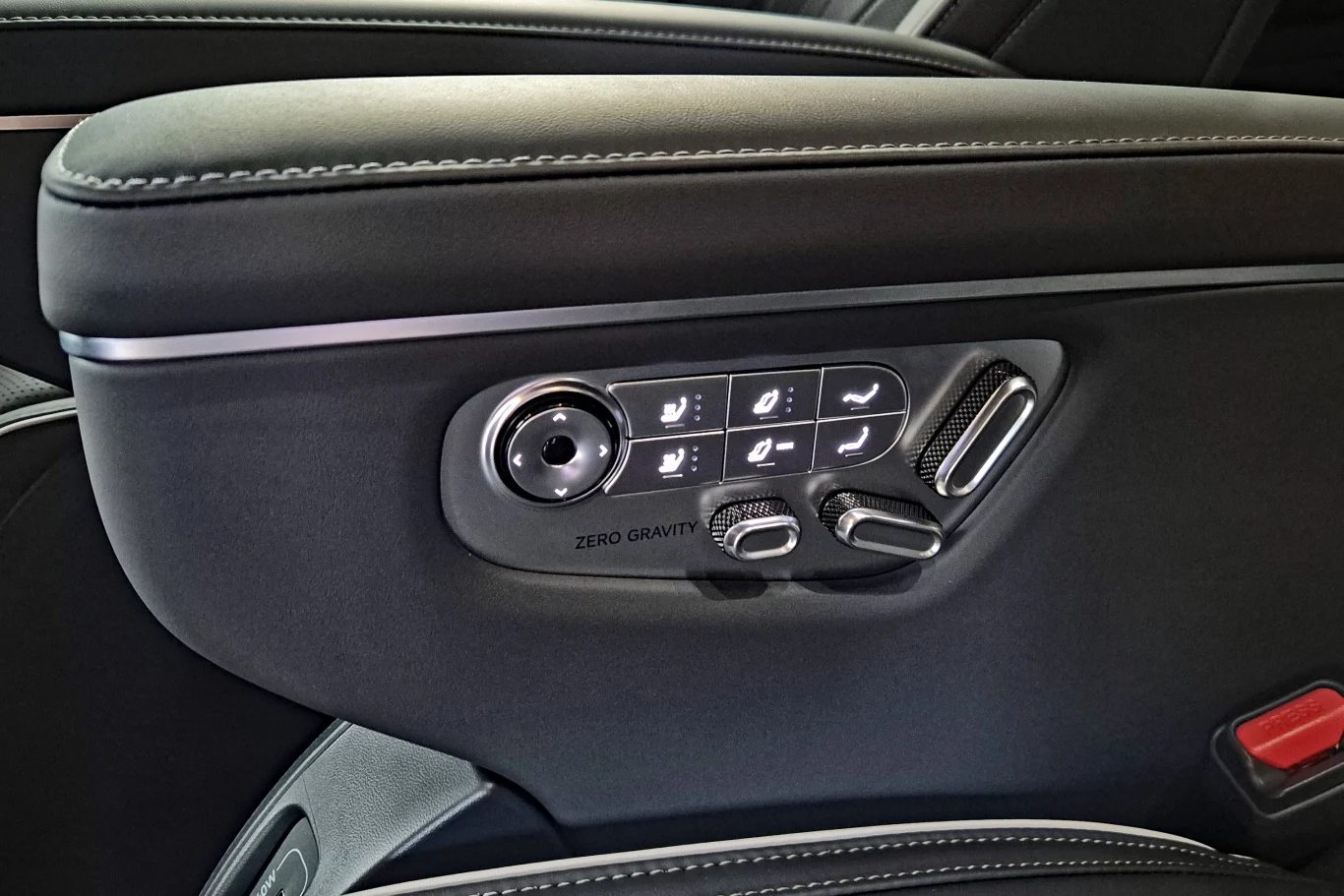 Panel sterowania elektrycznym fotelem samochodowym z przyciskami regulacji położenia, wsparcia lędźwiowego i funkcji Zero Gravity, otoczony czarną tapicerką z białymi przeszyciami.