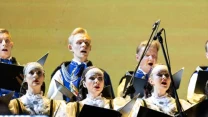 Chórzyści w ludowych strojach śpiewają na scenie, trzymając nuty w rękach, na tle jasnego, jednolitego ekranu.