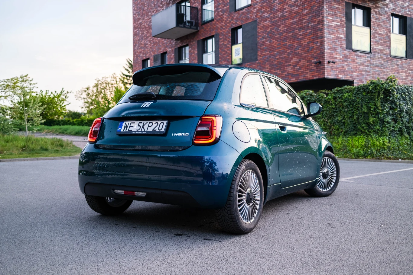 Zielony samochód Fiat 500 Hybrid zaparkowany na asfaltowym parkingu obok nowoczesnego budynku z cegły.