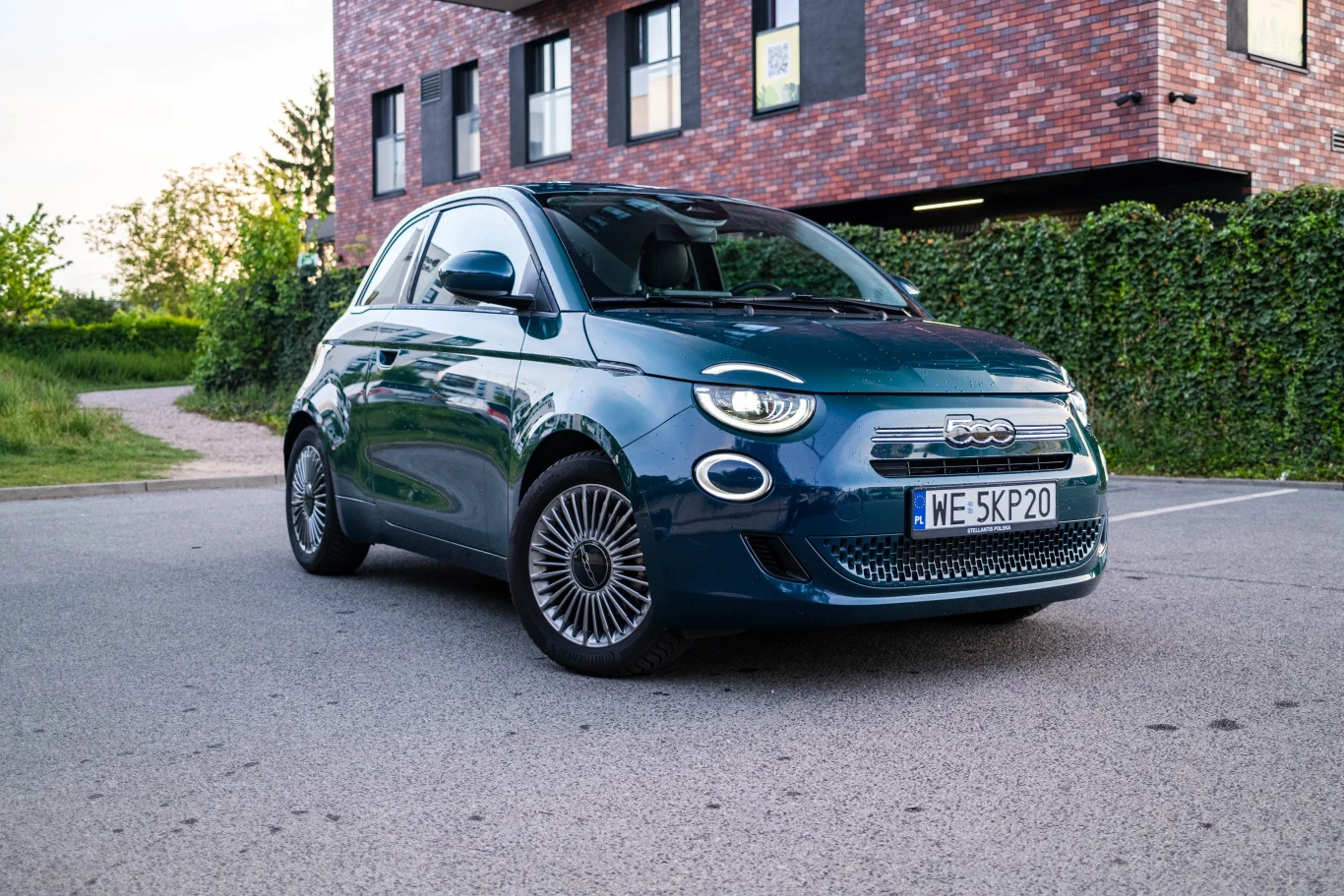Niebieski Fiat 500 zaparkowany na asfaltowej nawierzchni przed nowoczesnym budynkiem z cegły, w otoczeniu zieleni.