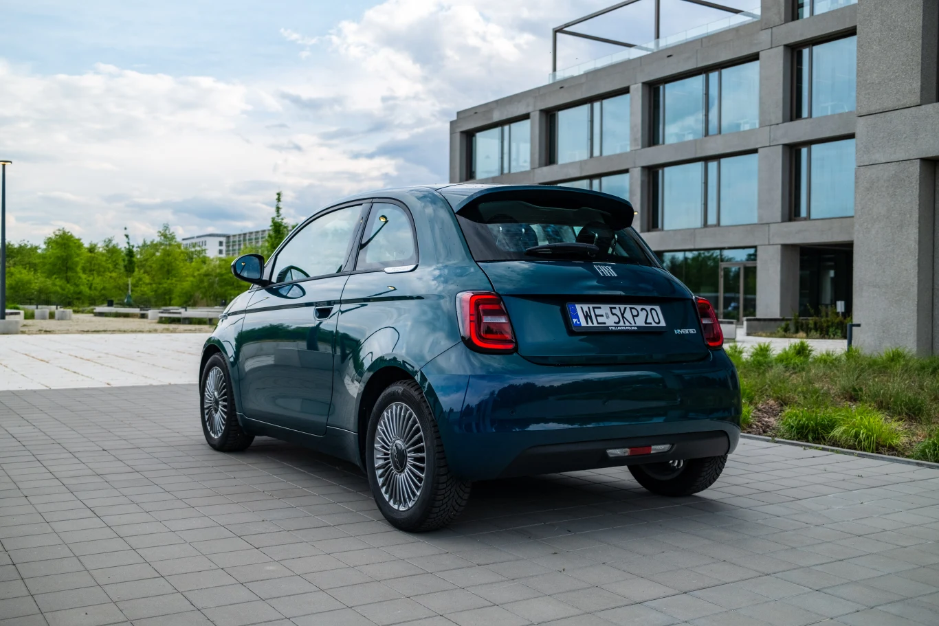 Niebieski samochód osobowy Fiat 500 zaparkowany na kostce brukowej w pobliżu nowoczesnego, przeszklonego budynku.