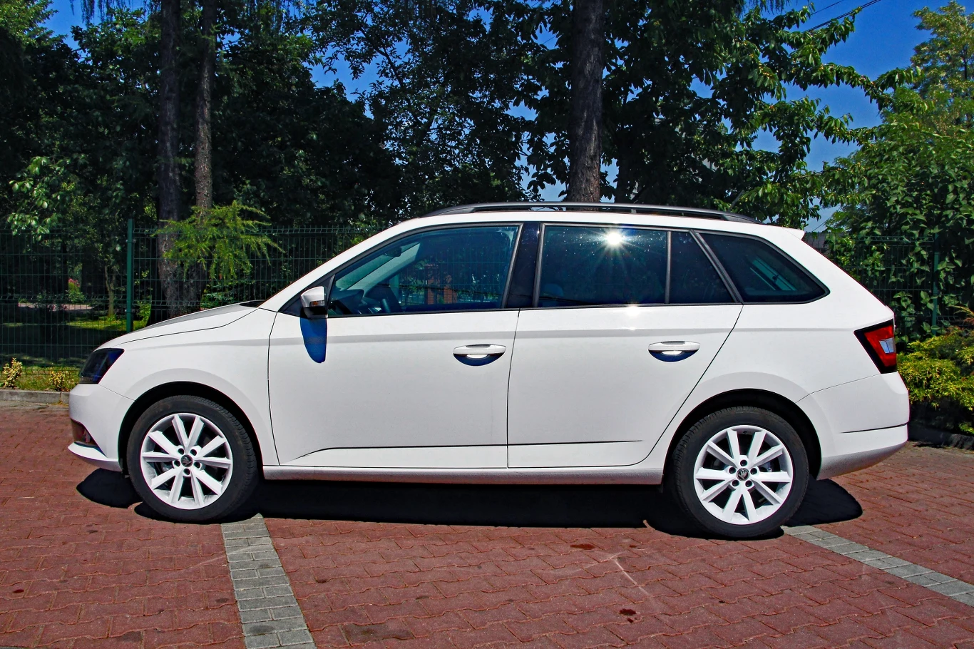 Skoda Fabia III kombi Biały samochód typu kombi zaparkowany na wyznaczonym miejscu, sfotografowany z boku, na tle zielonych drzew.
