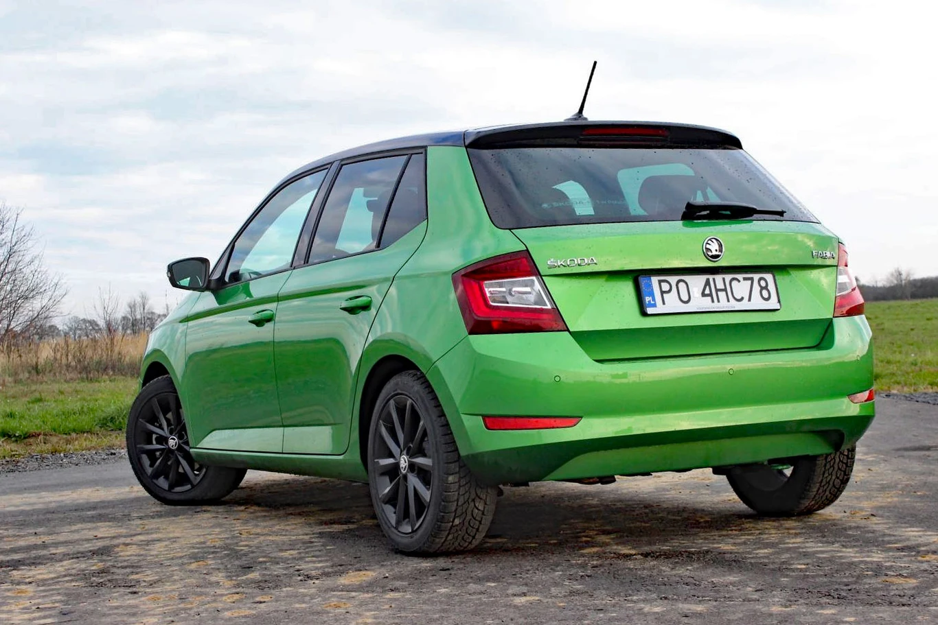 Skoda Fabia III Zielony hatchback Skoda Fabia z czarnymi felgami zaparkowany na betonowej nawierzchni przy polu, widok od tyłu po skosie.