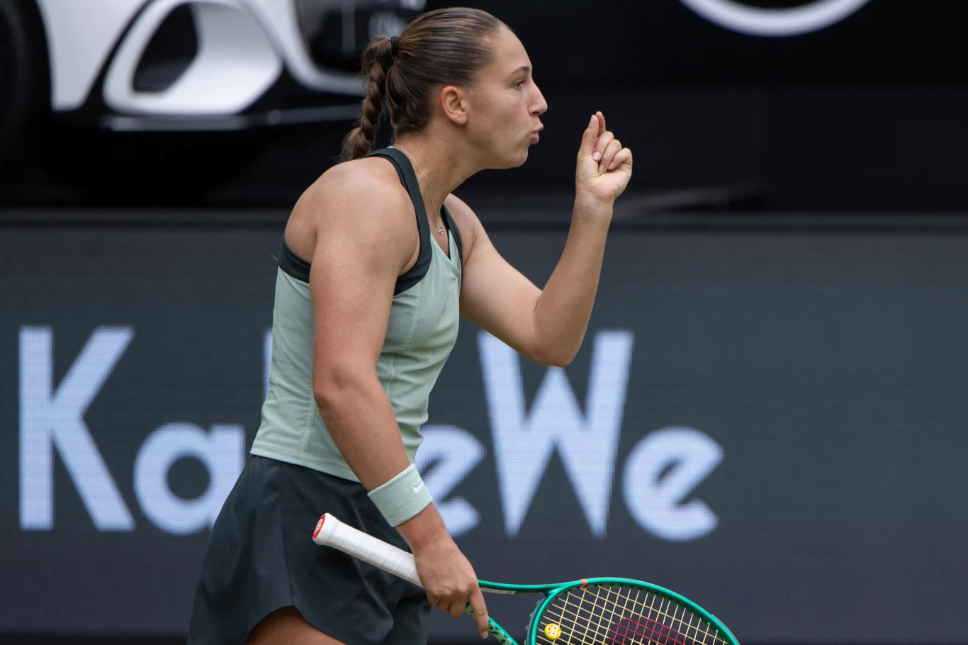 Tenisistka w sportowym stroju z uniesionym palcem wskazującym wykonuje gest, trzymając rakietę do tenisa w lewej dłoni na tle kortu i dużej reklamy.