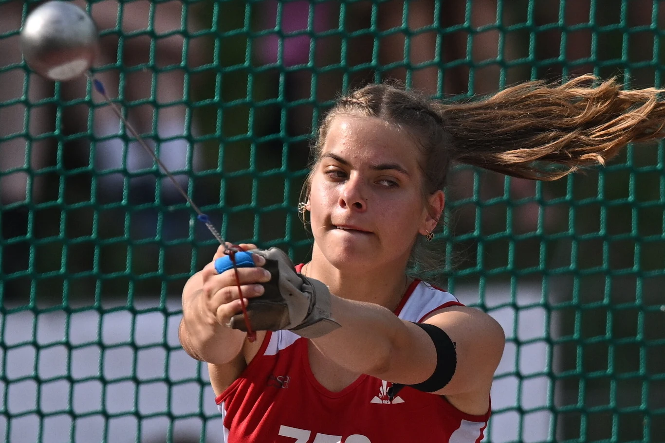 Zawodniczka w czerwonym stroju sportowym wykonuje rzut młotem, trzymając rączkę młota oburącz i koncentrując się na ruchu, w tle widoczne zielone ogrodzenie zabezpieczające.
