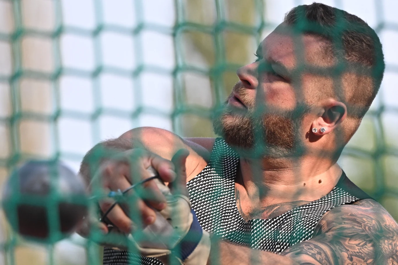 Atleta w trakcie rzutu młotem, skupiony wyraz twarzy, umięśnione ramiona z widocznym tatuażem i siatka ochronna na pierwszym planie.