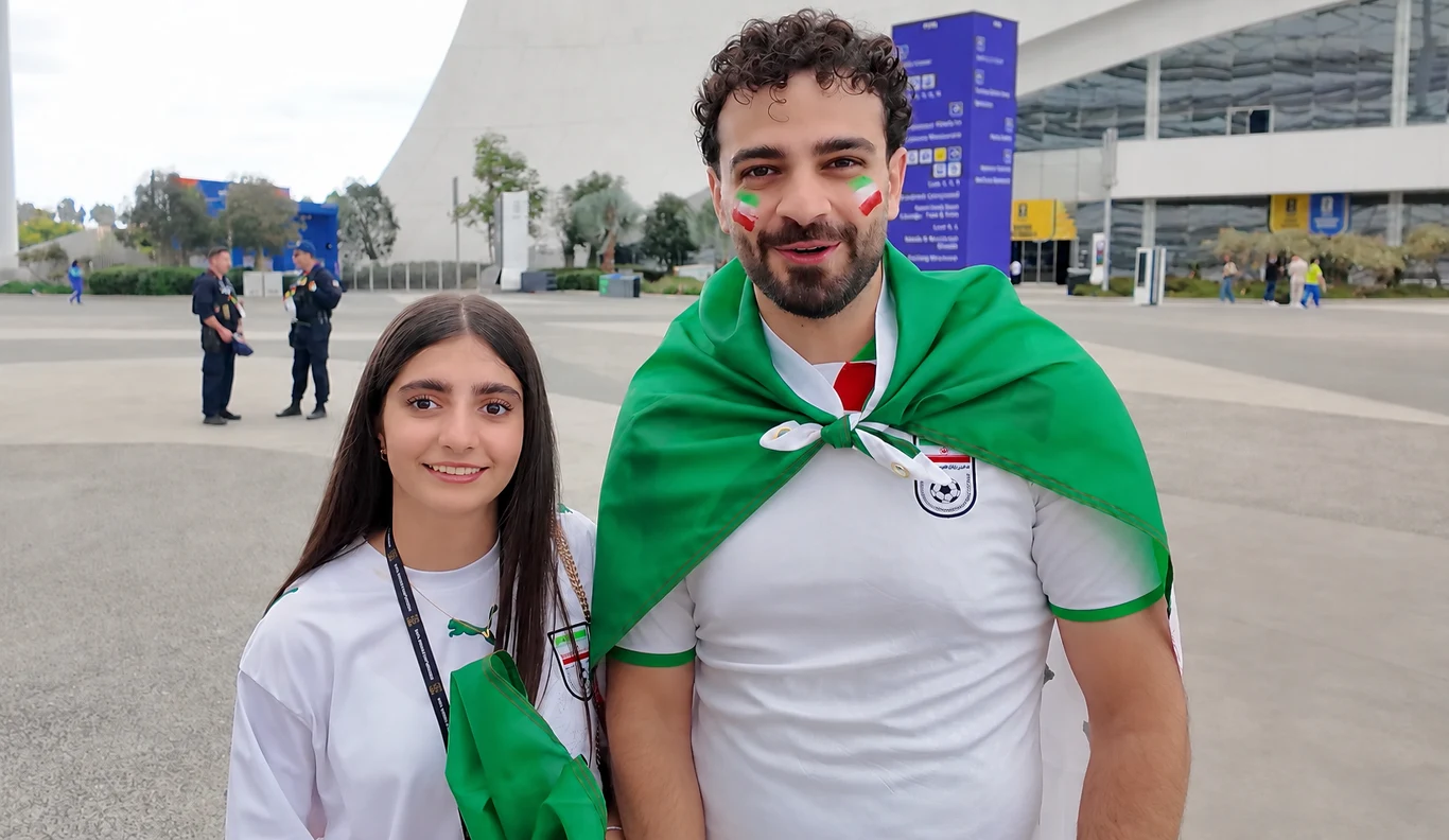 Kibice w białych koszulkach z flagami i malowidłami w barwach narodowych na twarzach, jeden z nich z zieloną flagą zarzuconą na ramiona, w tle nowoczesny stadion.